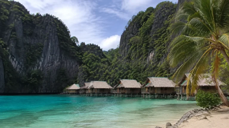 Thatched bungalows beside turquoise water beneath steep limestone cliffs.