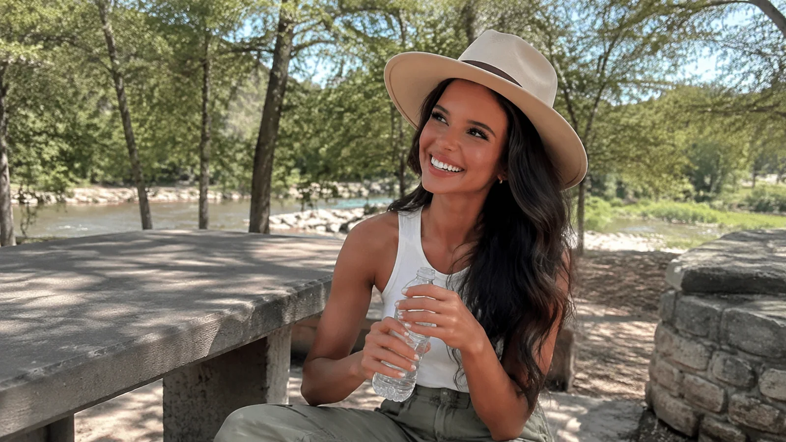Woman wearing a wide-brimmed hat holding a water bottle at a shaded picnic table.