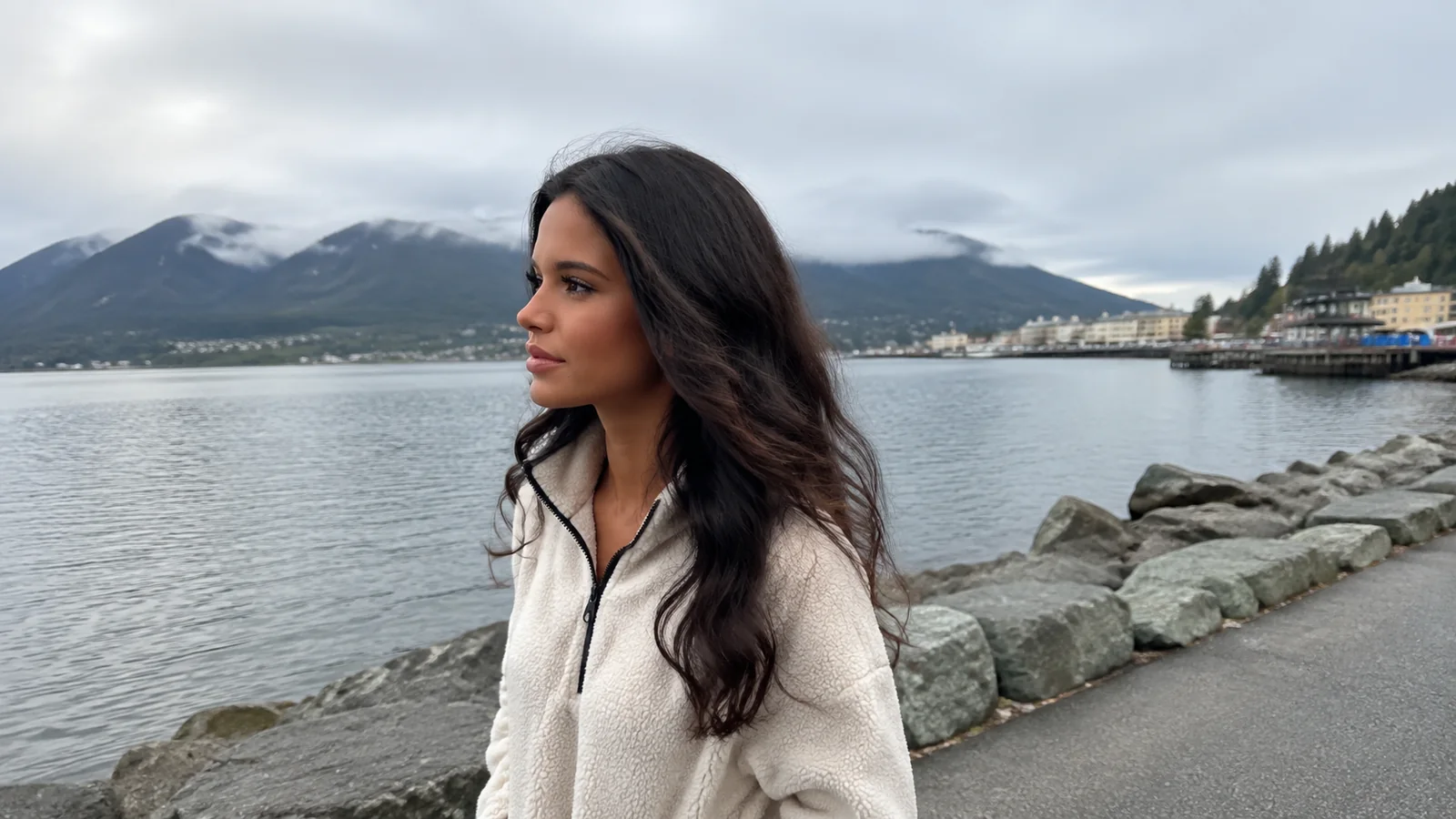 Woman walking beside a rocky waterfront with cloud-covered mountains across the bay.