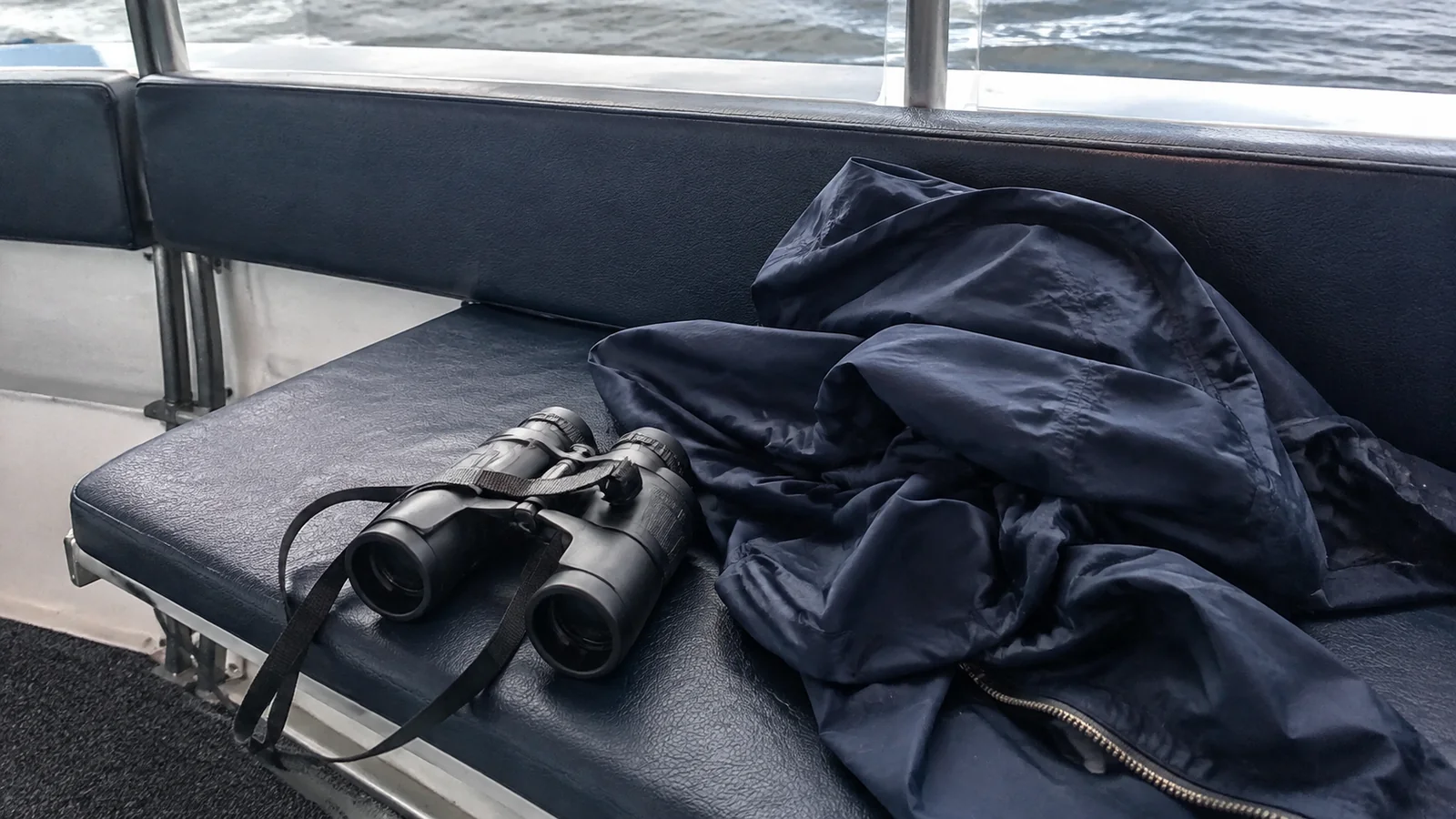Binoculars and a rain jacket on a seat beside a ship window.