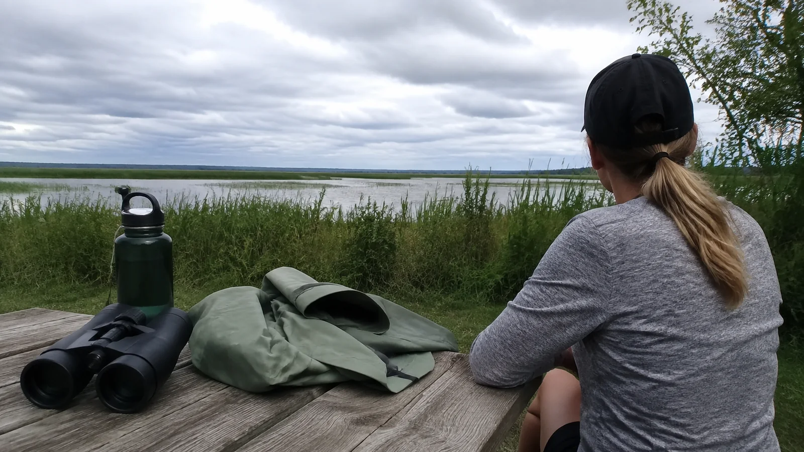 Woman at a lakeside picnic table with binoculars, a water bottle, and a jacket.