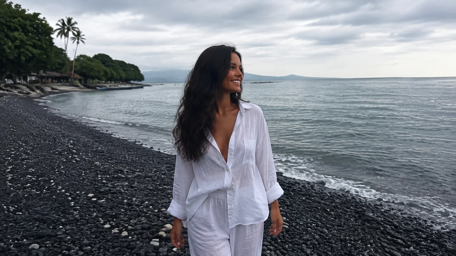 Woman stands on a black pebble beach beside calm water.