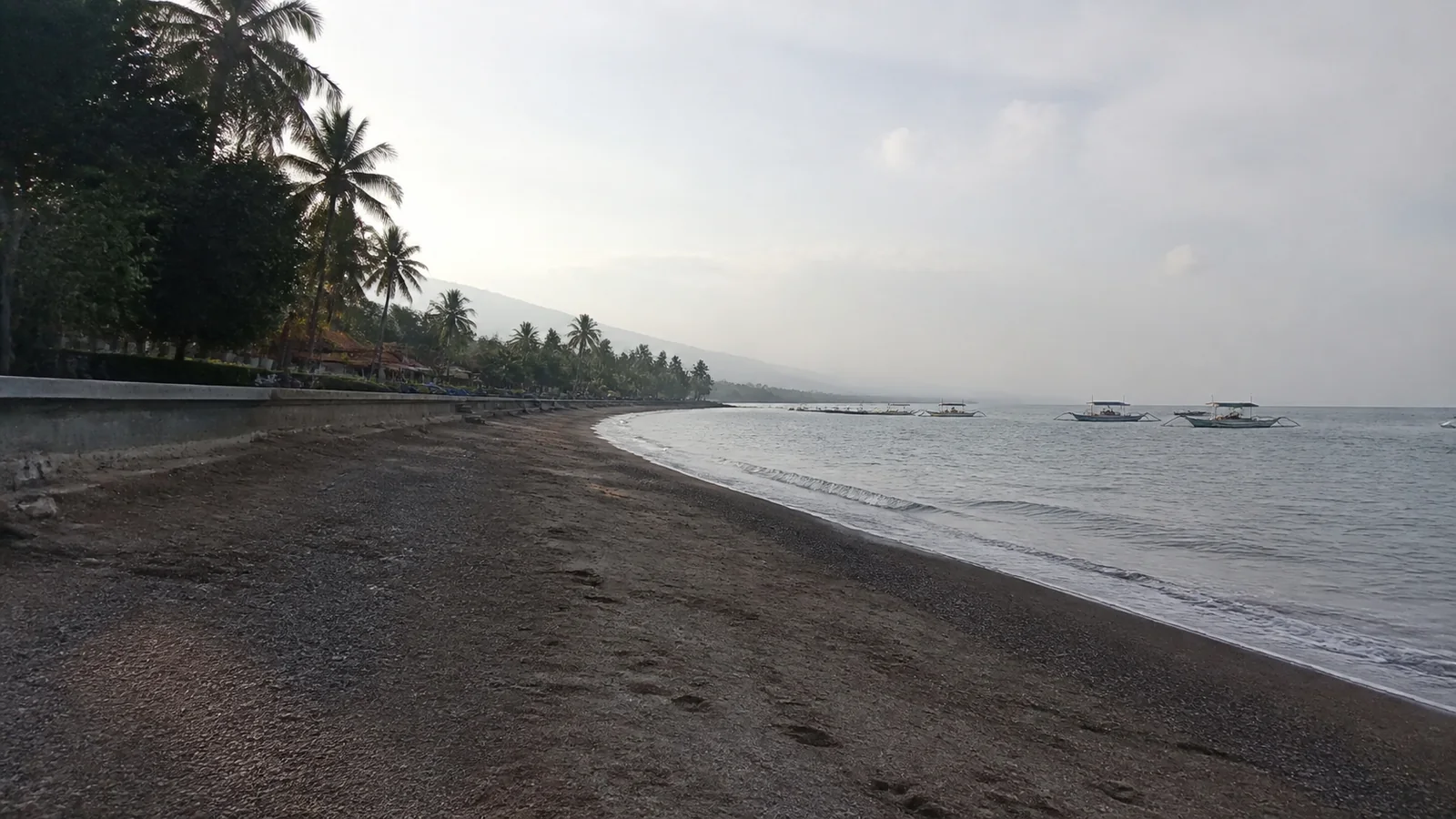 Dark sandy shoreline curves beneath palms toward hazy coastal hills.