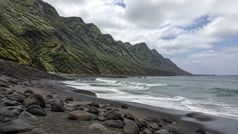 Black sand and boulders border the sea beneath steep green mountains.