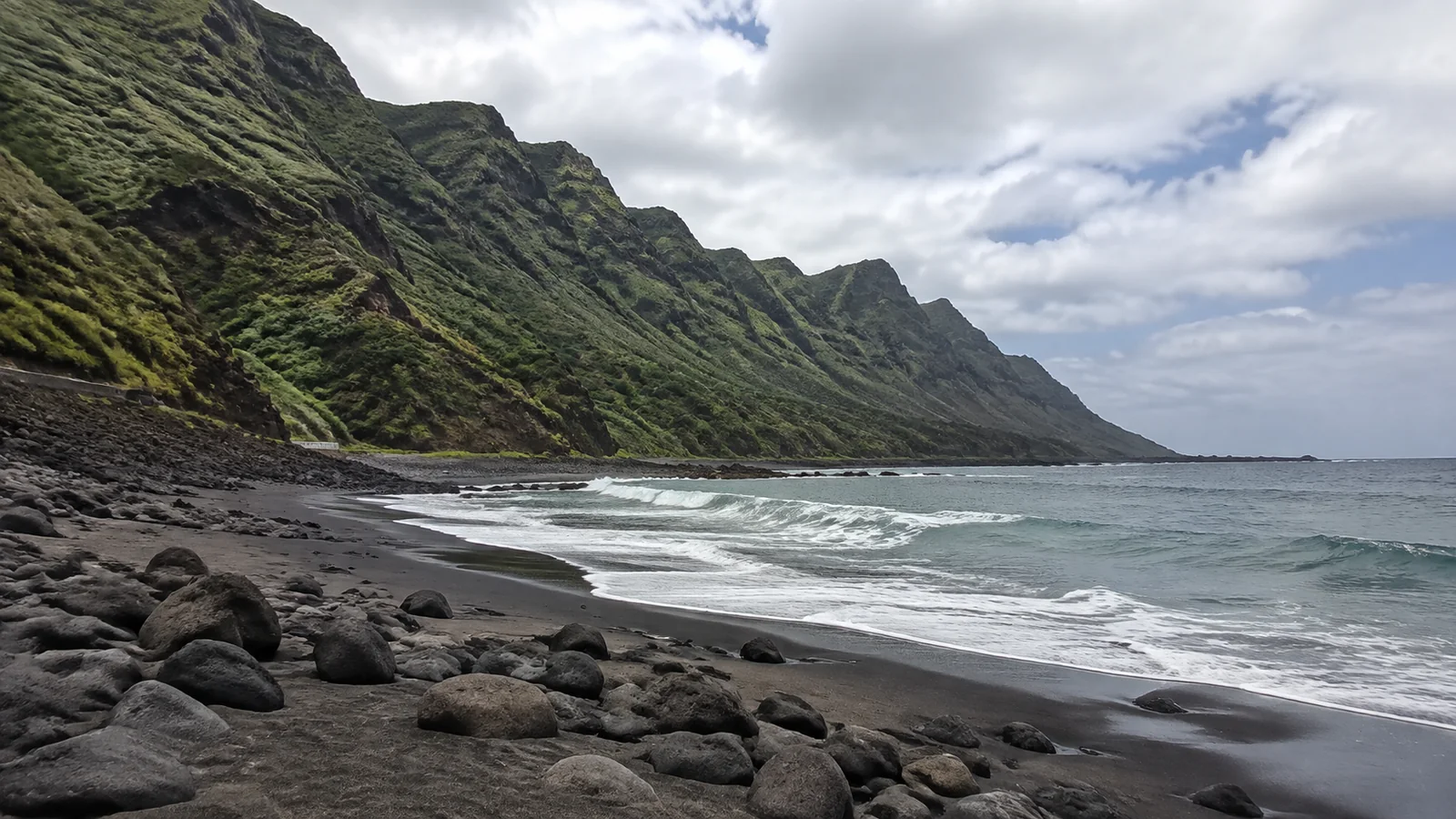 Black sand and boulders border the sea beneath steep green mountains.