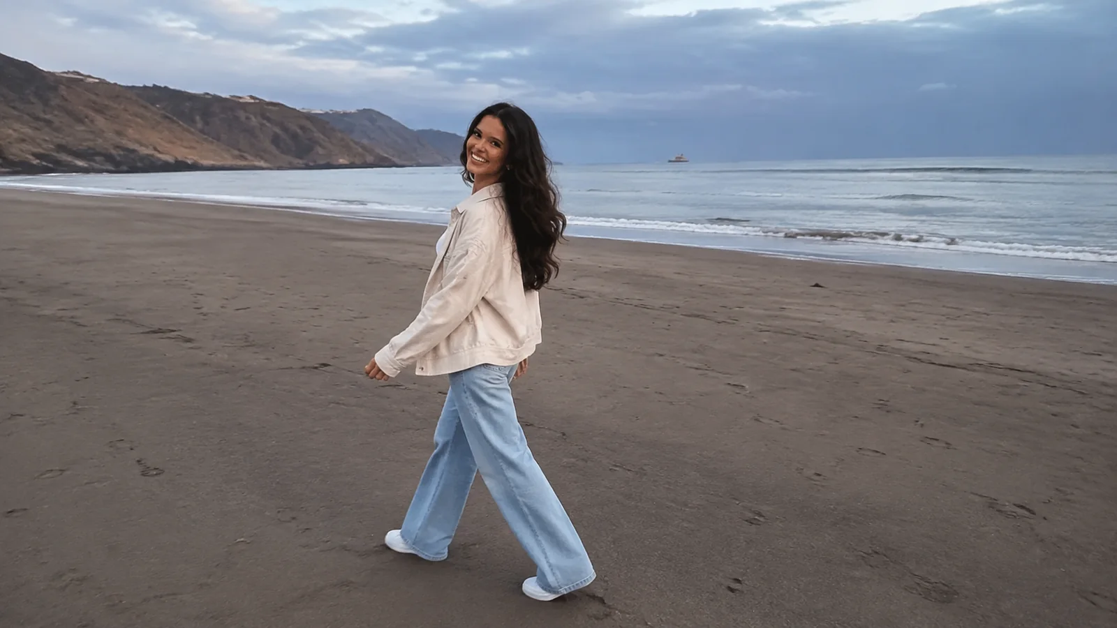 Woman walks along a broad dark sandy beach.