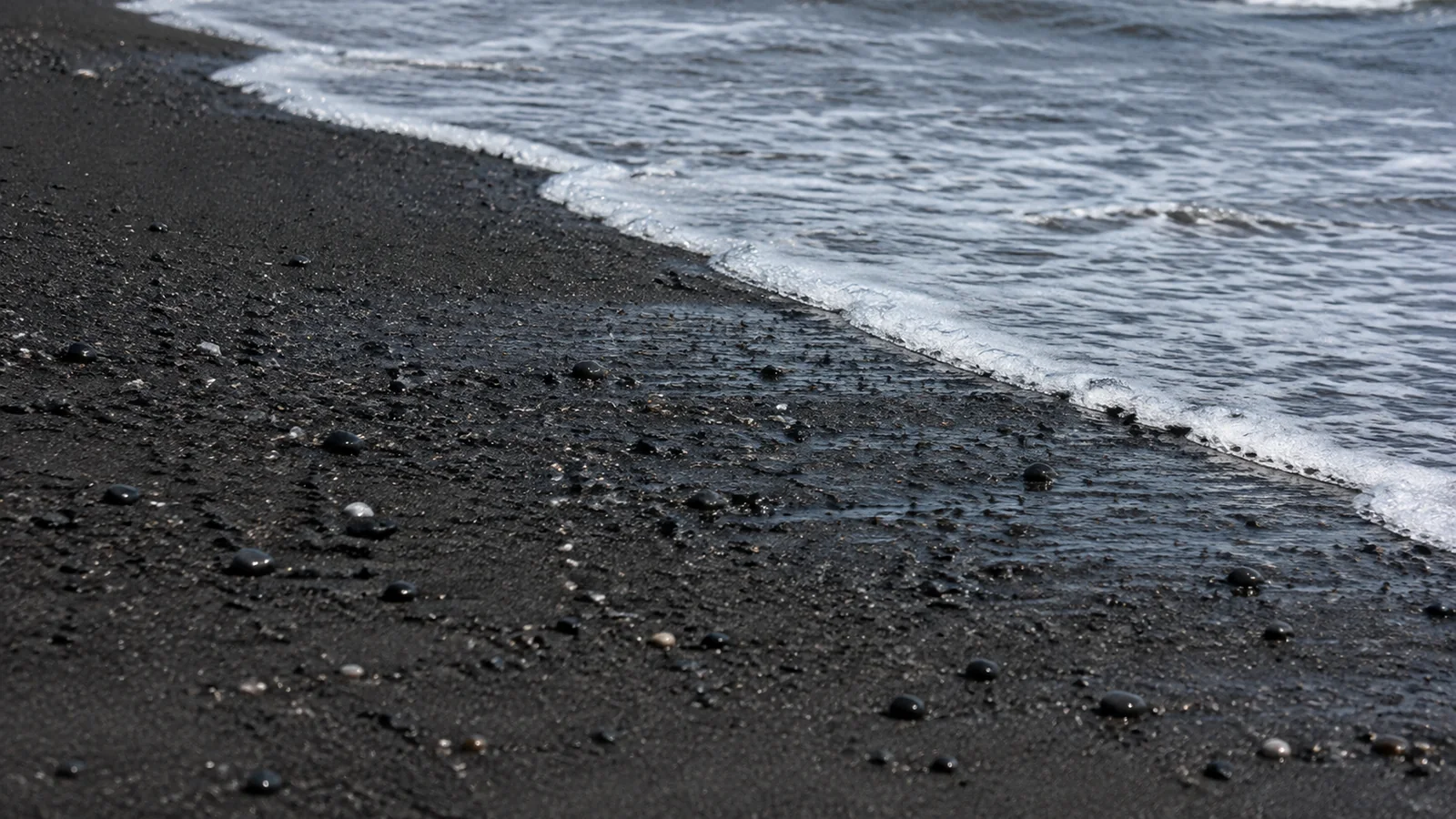Foamy wave washes over dark pebbles and black sand.