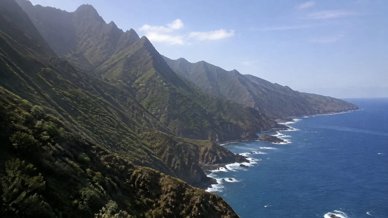 Jagged green coastal ridges descend sharply into deep blue water.