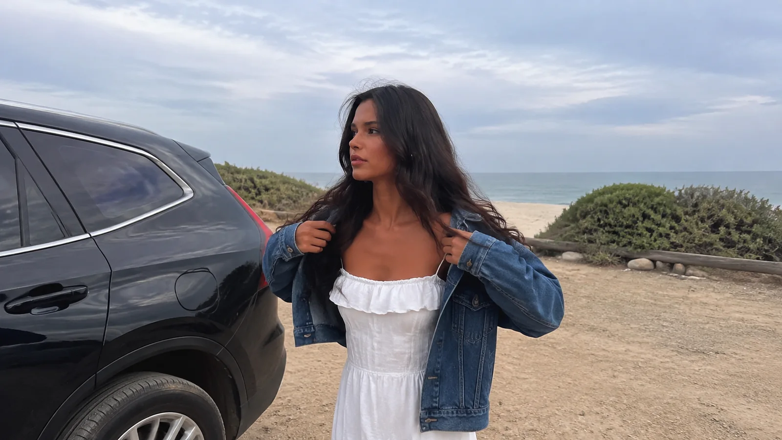 Woman adjusting her denim jacket beside a car near the beach.