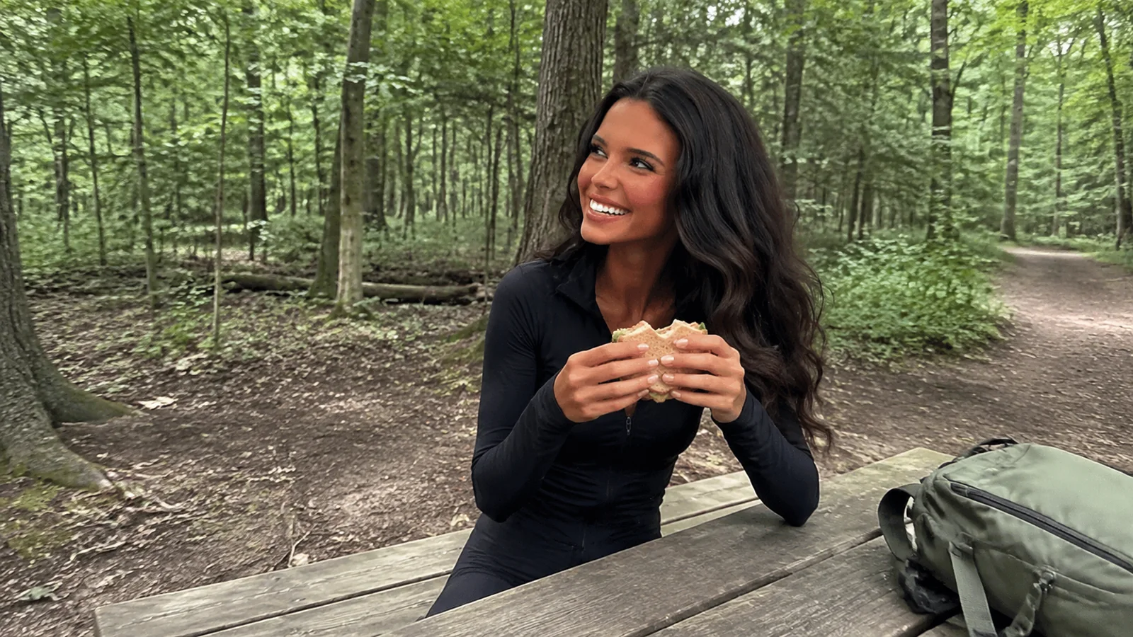 Woman eating a sandwich at a forest picnic table beside a backpack.