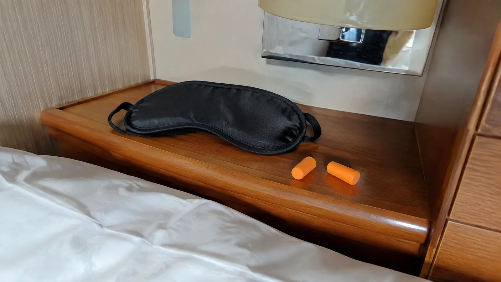 Sleep mask and orange earplugs sit on a wooden bedside shelf.
