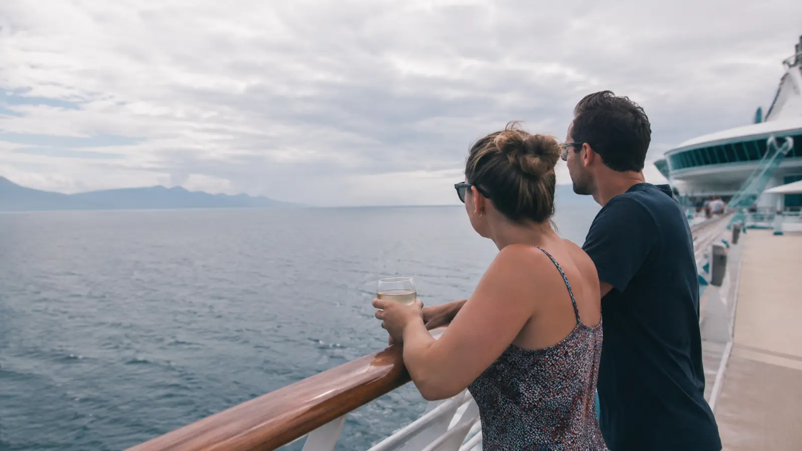 Couple standing at a cruise ship railing while one holds a wine glass