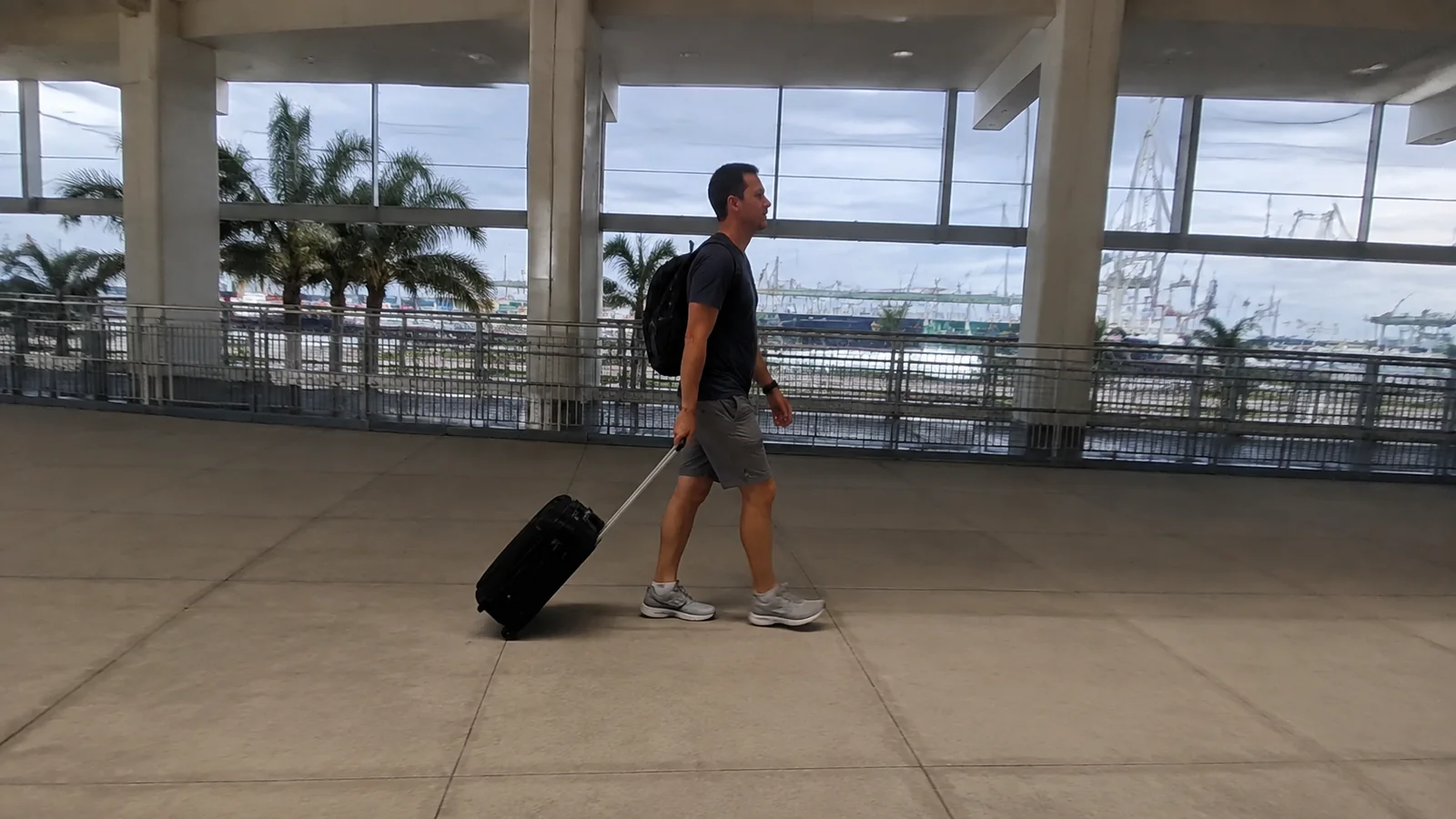 Man pulls a wheeled suitcase through a bright waterfront terminal.