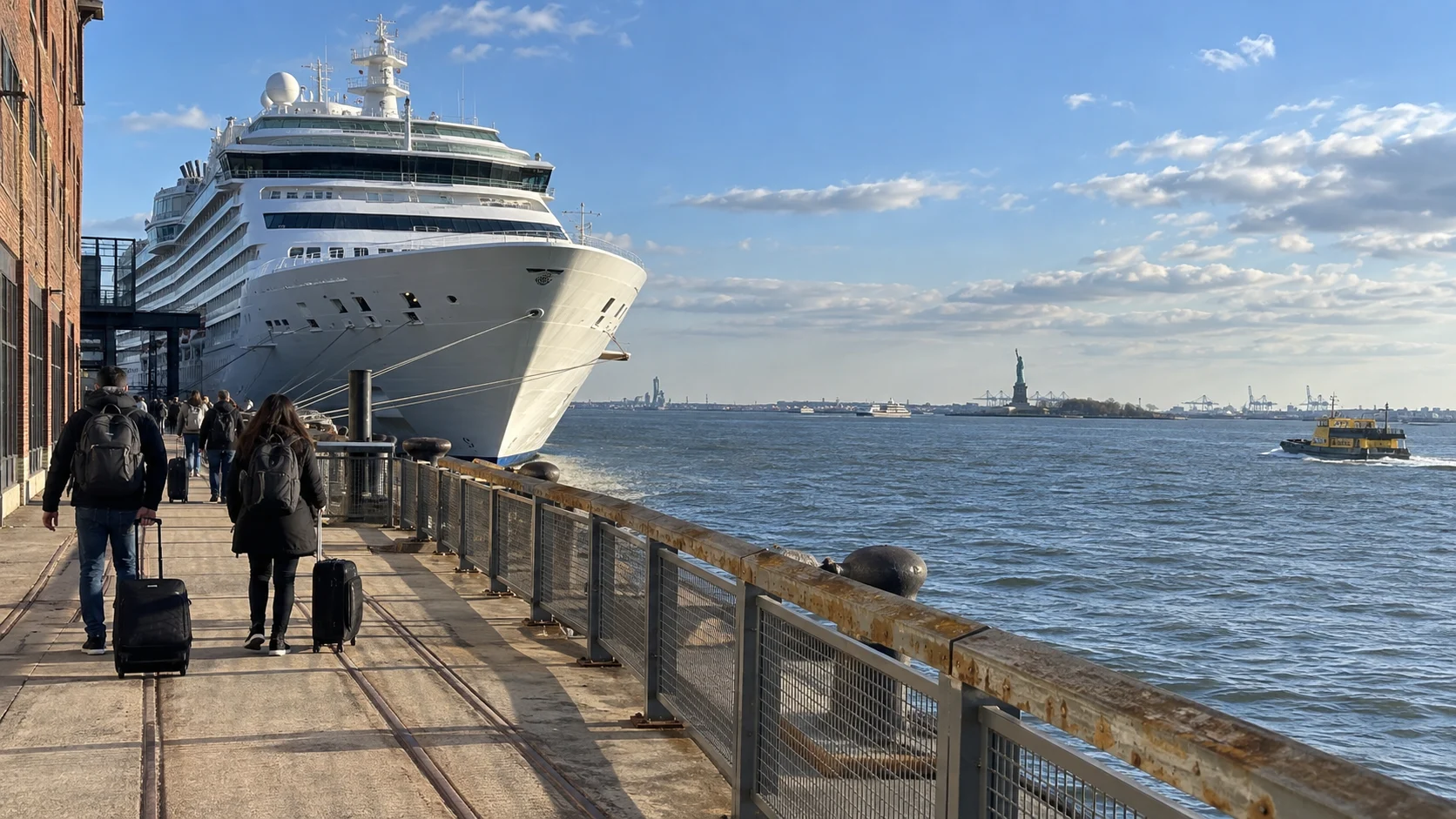 Travelers walking toward a cruise ship at Brooklyn Cruise Terminal with the Statue of Liberty across the harbor.