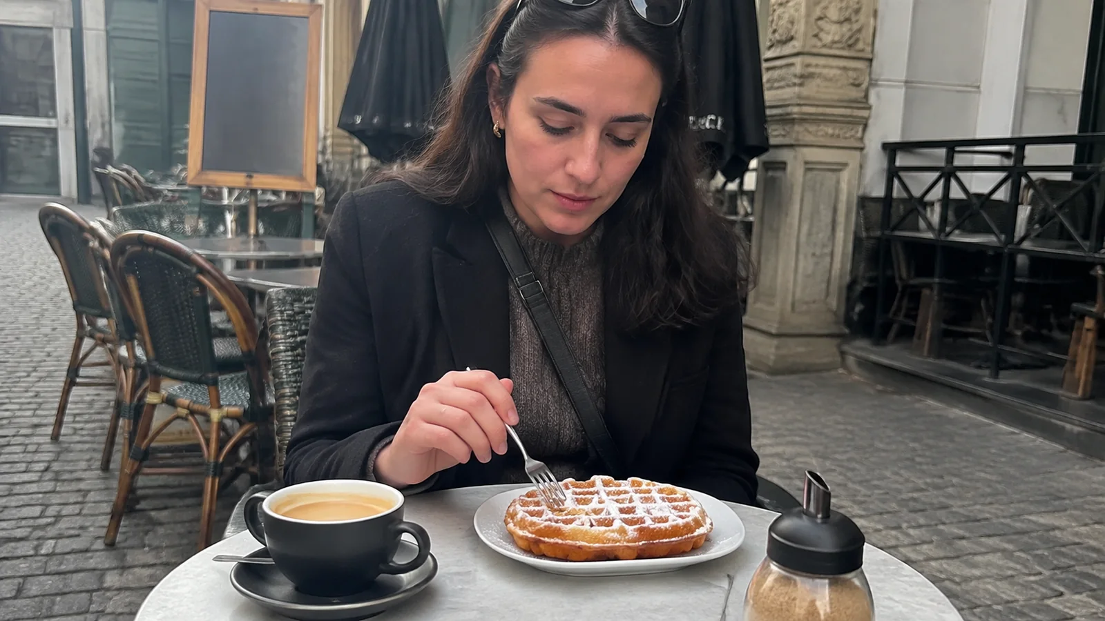 Woman eating a powdered waffle beside coffee at an outdoor cafe.