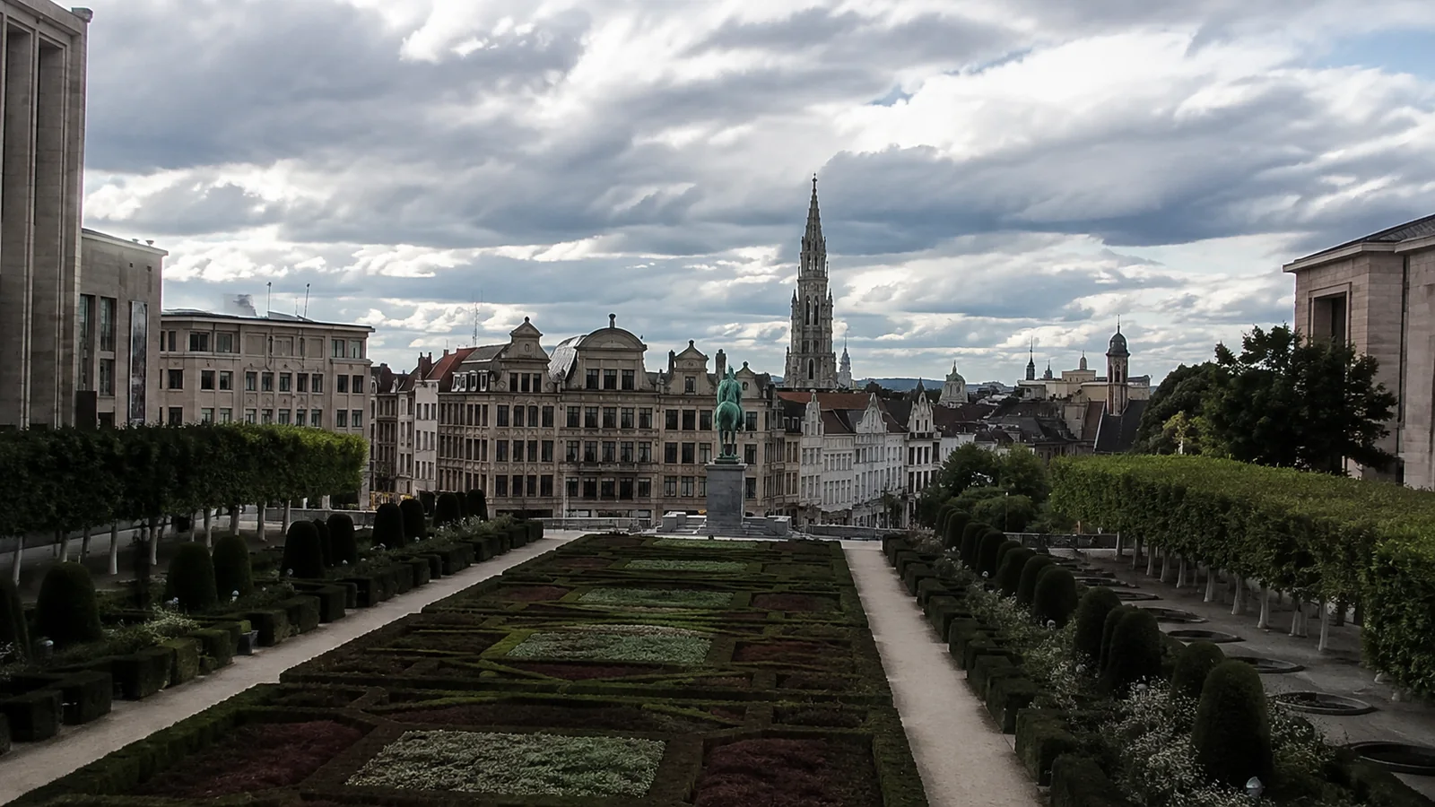 Formal geometric gardens below ornate buildings and a tall spire.