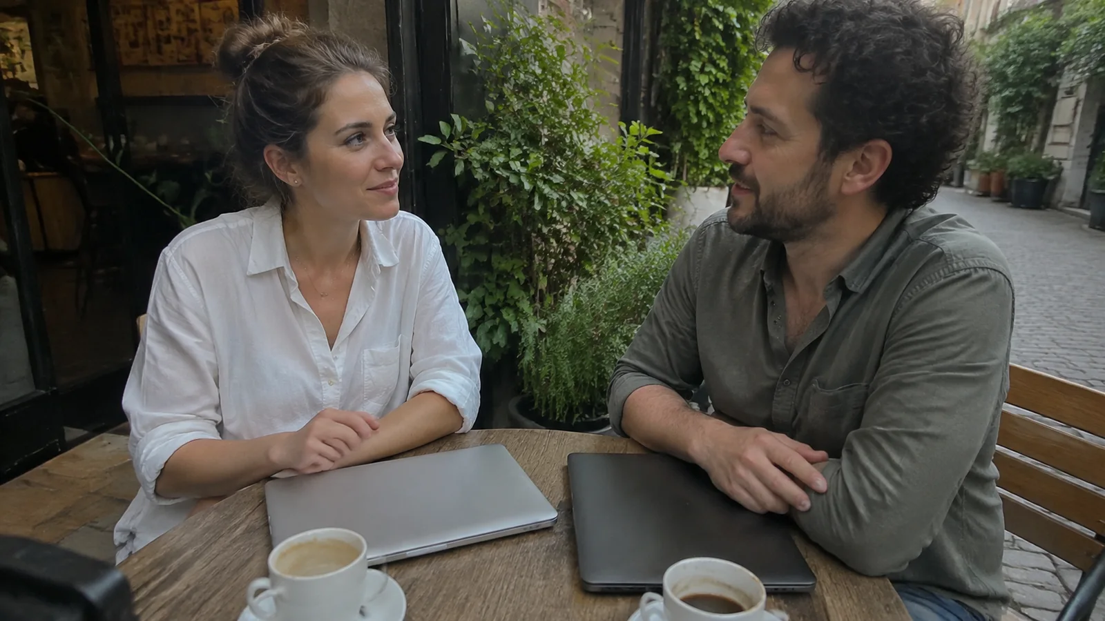Two people talking across an outdoor table with a closed laptop.