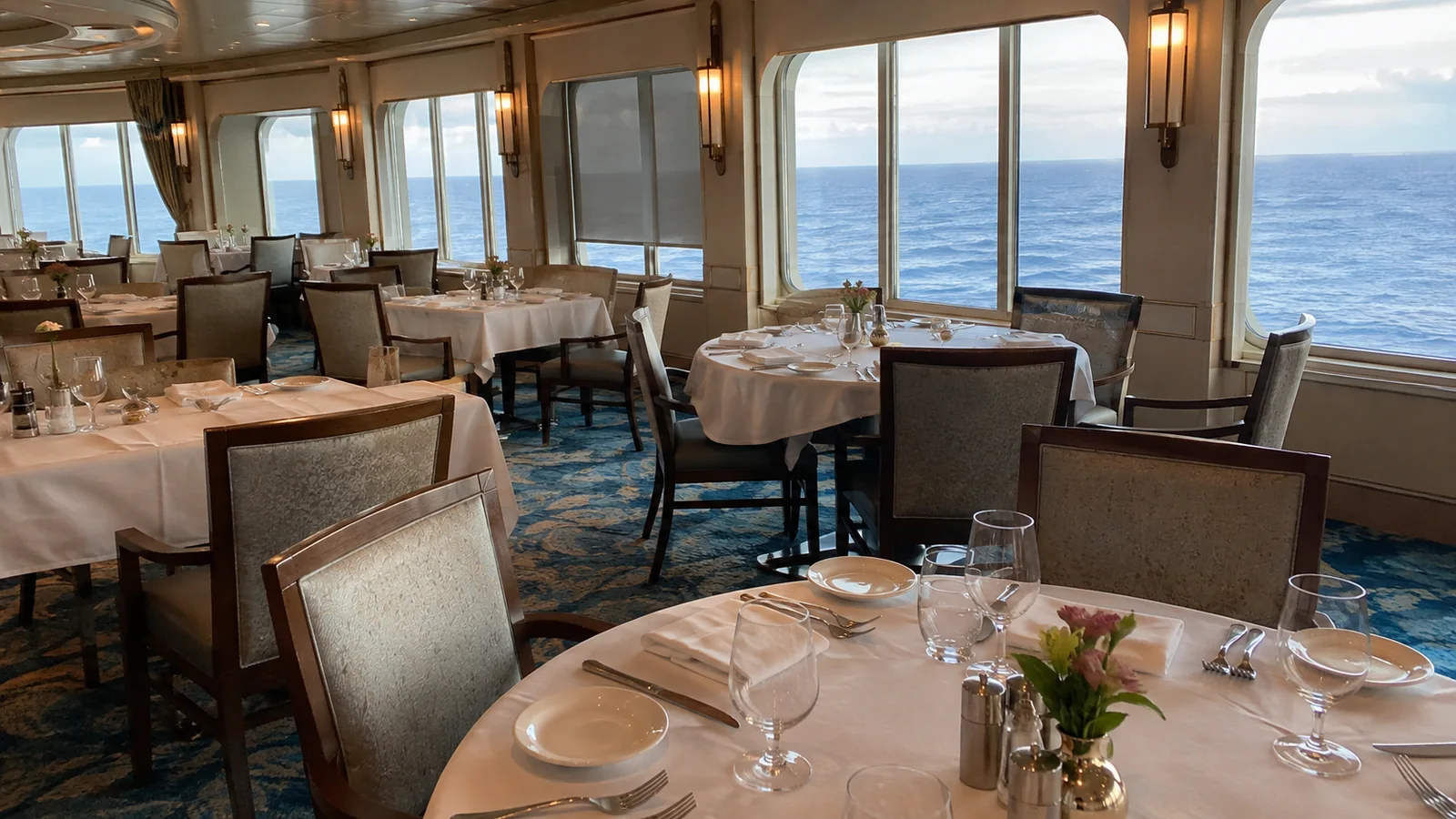 Set dining tables face large ocean windows in a ship restaurant.