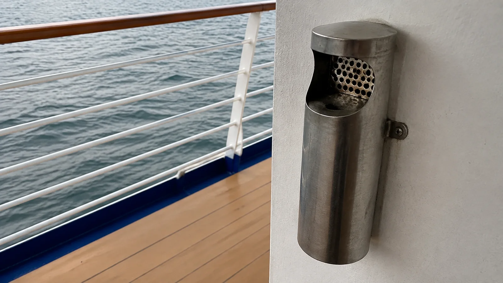 Wall-mounted metal cigarette receptacle beside an open deck railing.