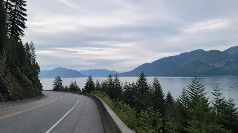 Road curving beside a mountain lake beneath a pale cloudy sky.