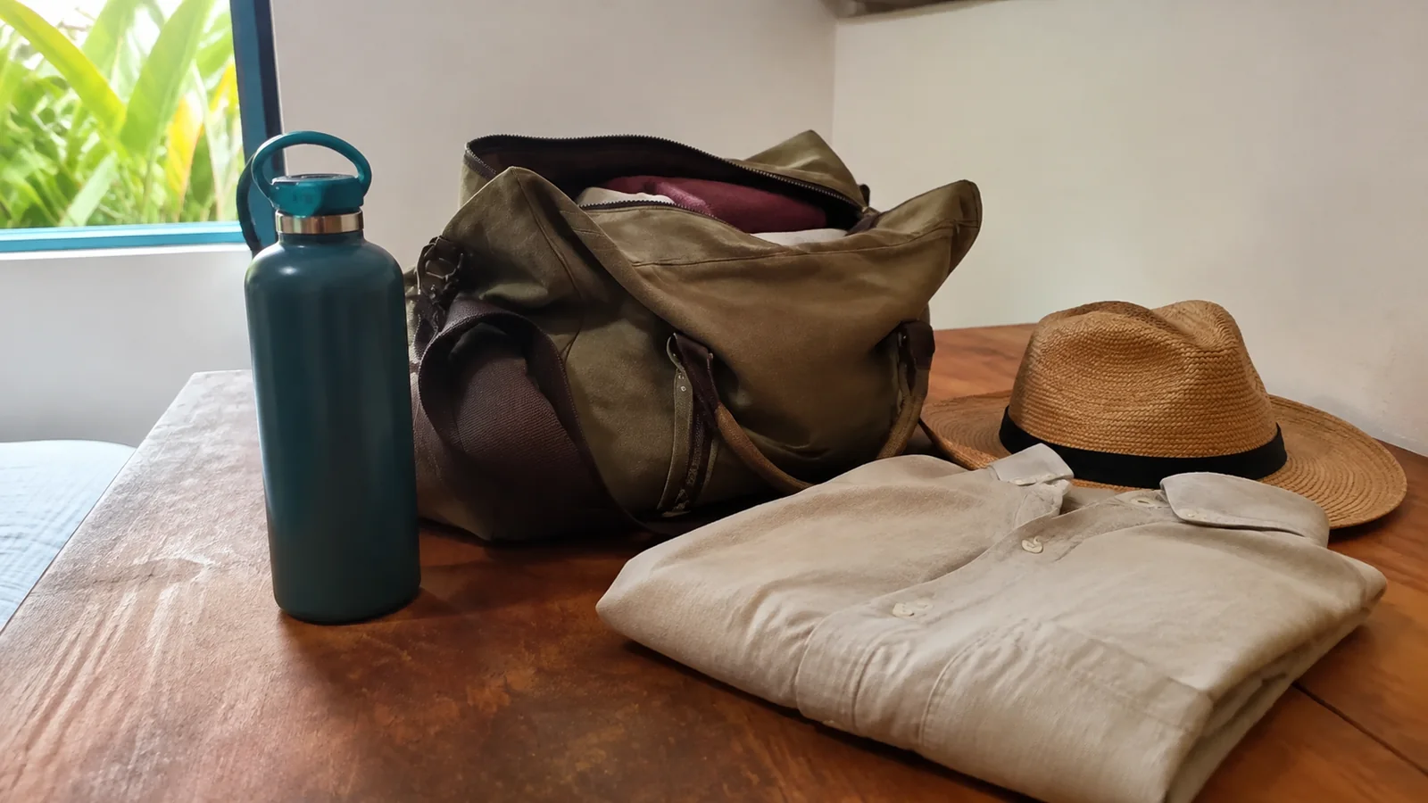 Travel bag, water bottle, folded shirt, and straw hat on a wooden table.