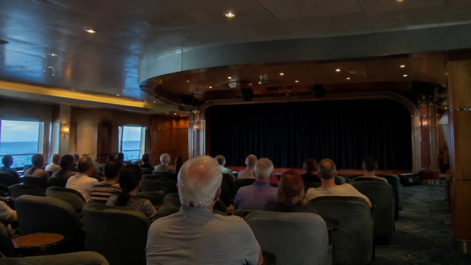 Seated audience faces a curtained stage in a ship lounge.