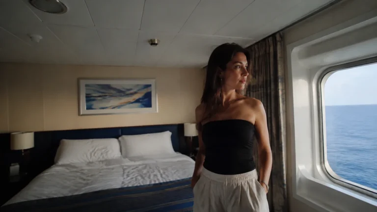 Woman stands beside a stateroom window overlooking the sea.