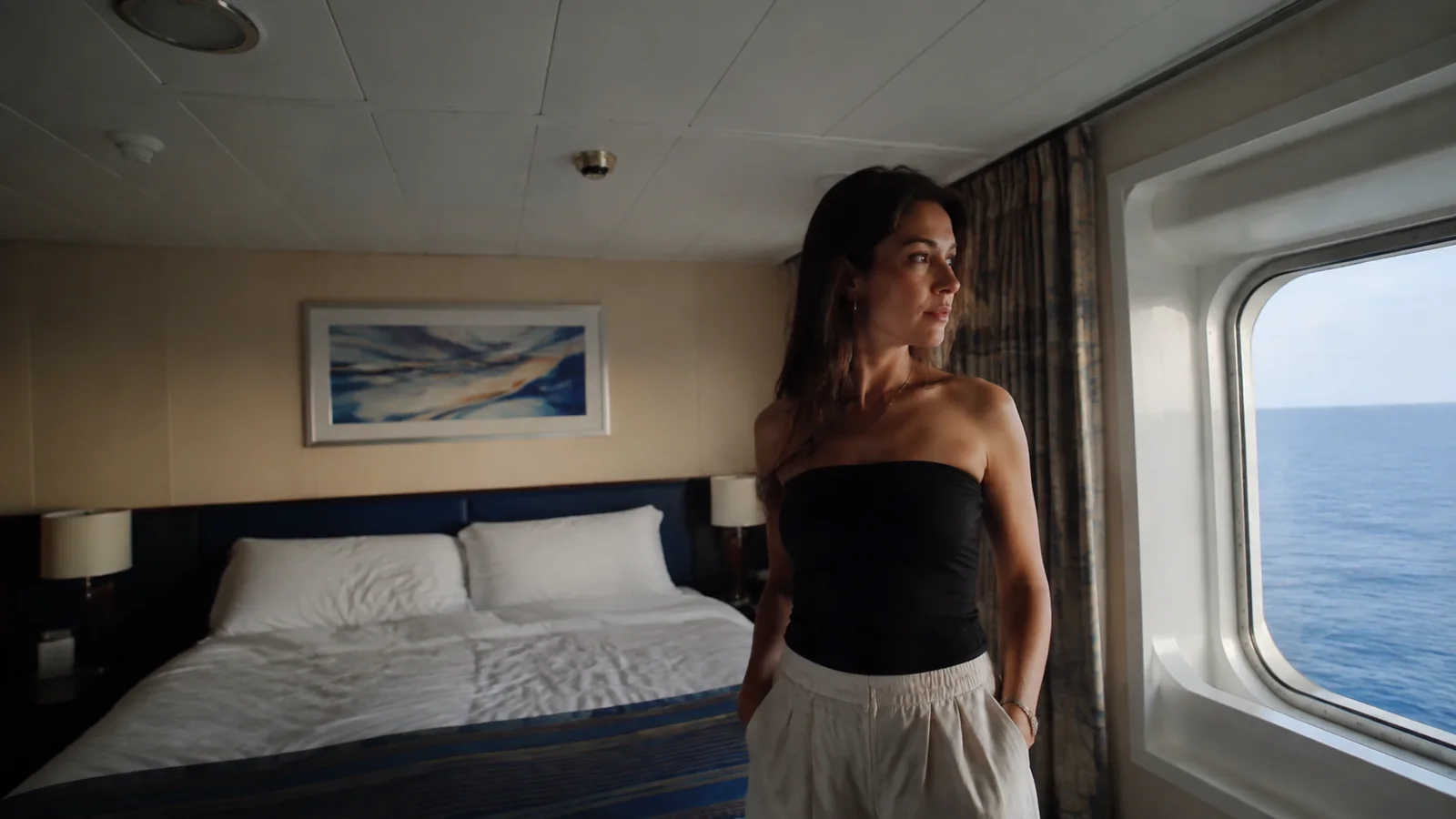 Woman stands beside a stateroom window overlooking the sea.