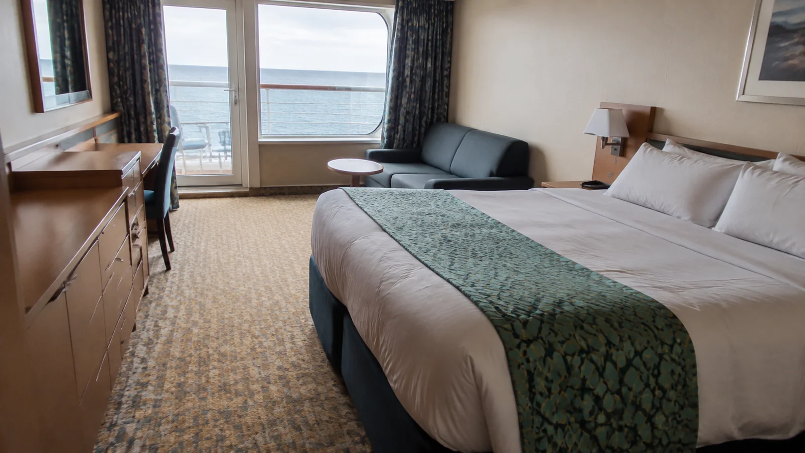Spacious ship cabin with a bed, sofa, and ocean-facing windows.