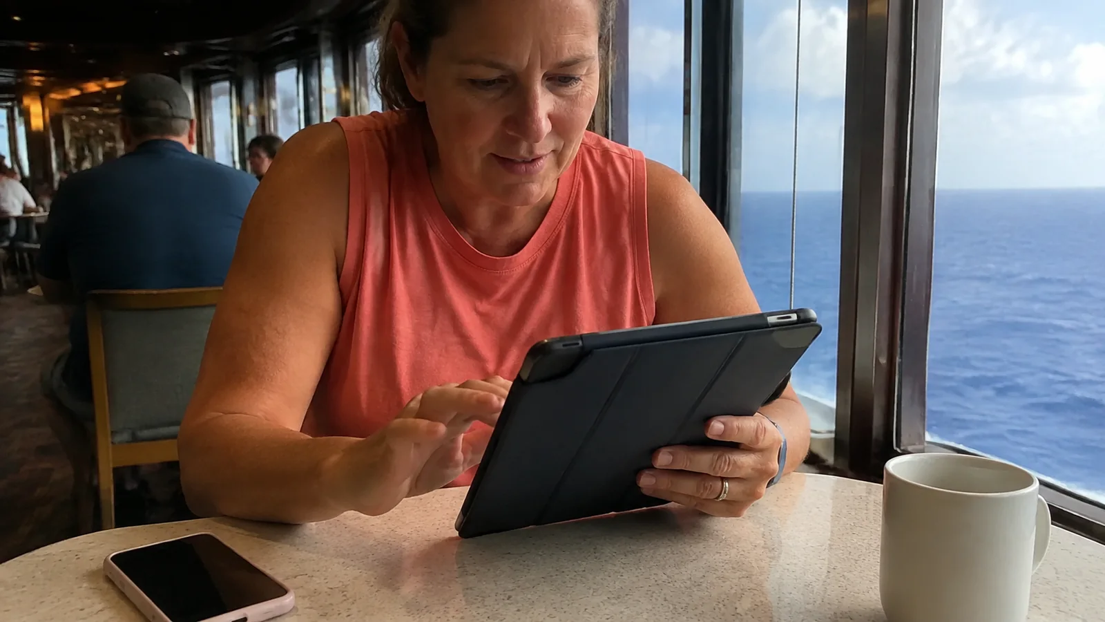 Passenger using a tablet beside a cup of coffee at a ship window.