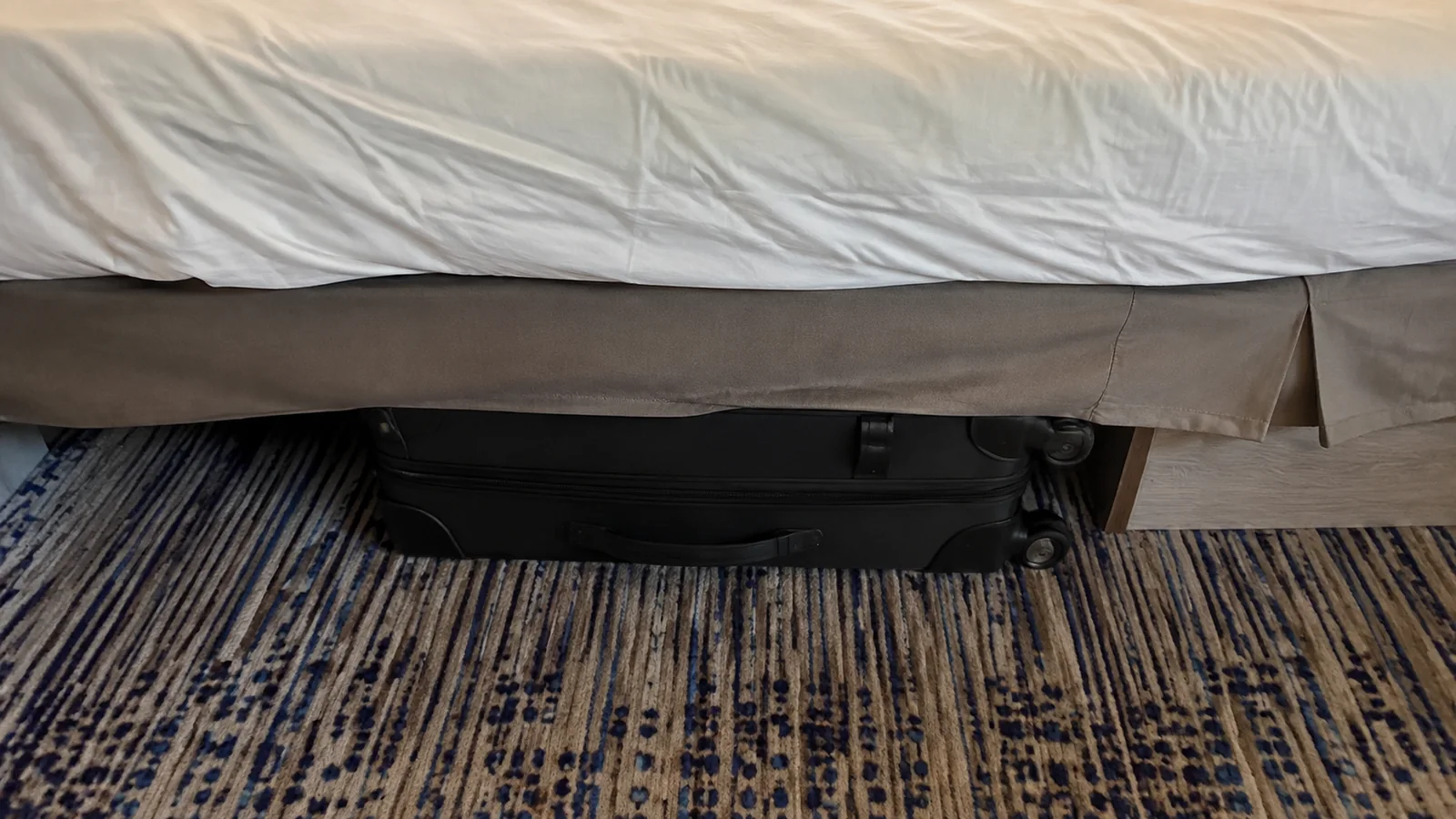 Black suitcase fits beneath a bed on patterned carpet.