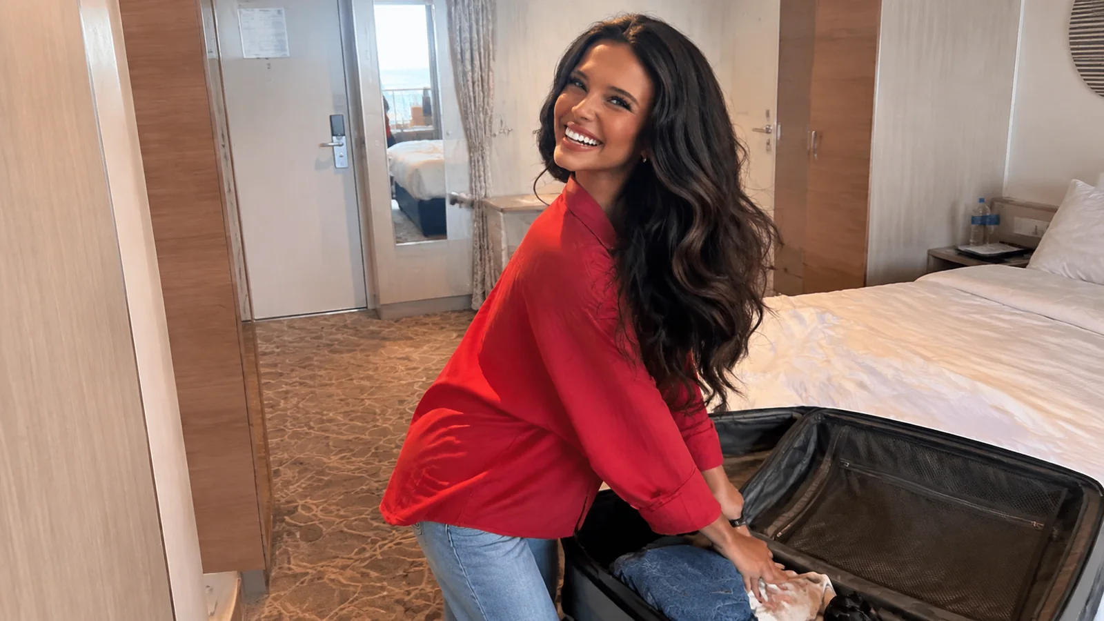 Woman packs an open suitcase on a bed while smiling.