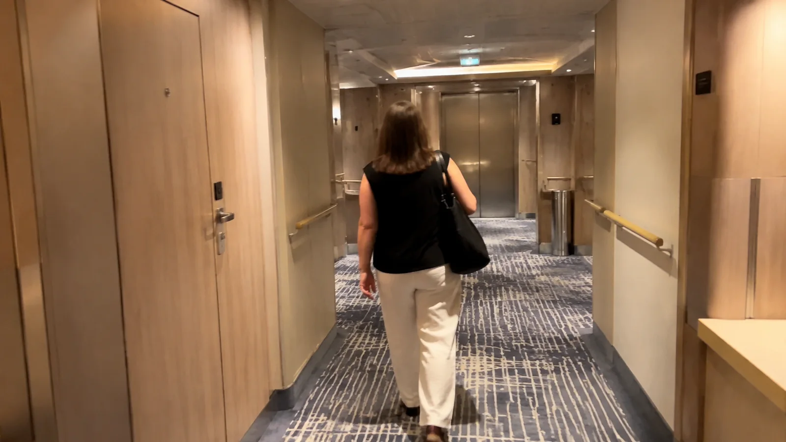 Woman walks toward elevators along a carpeted cruise ship corridor.
