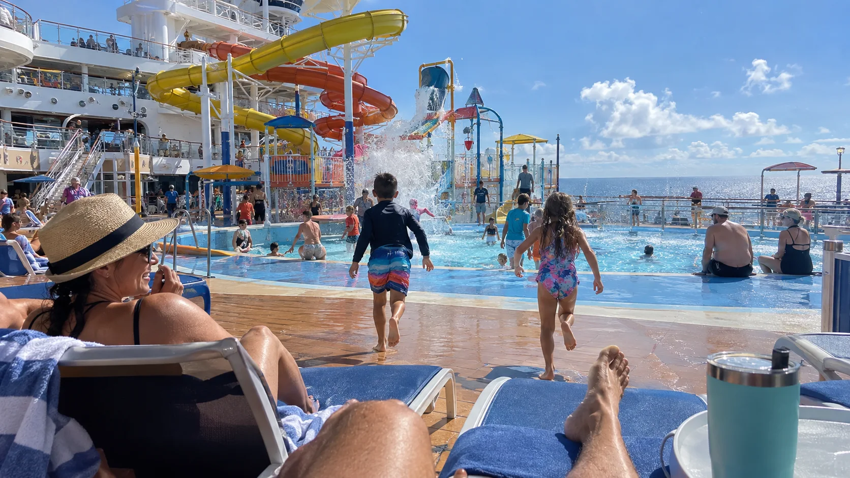 Family pool deck with waterslides and children running toward the splash area on a modern cruise ship.