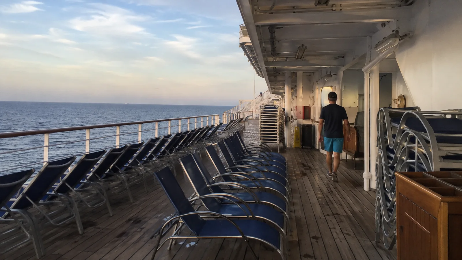 Passengers walking past lounge chairs on a cruise ship promenade at sunset