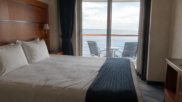 Cruise cabin with a double bed and two chairs on the ocean-facing balcony.