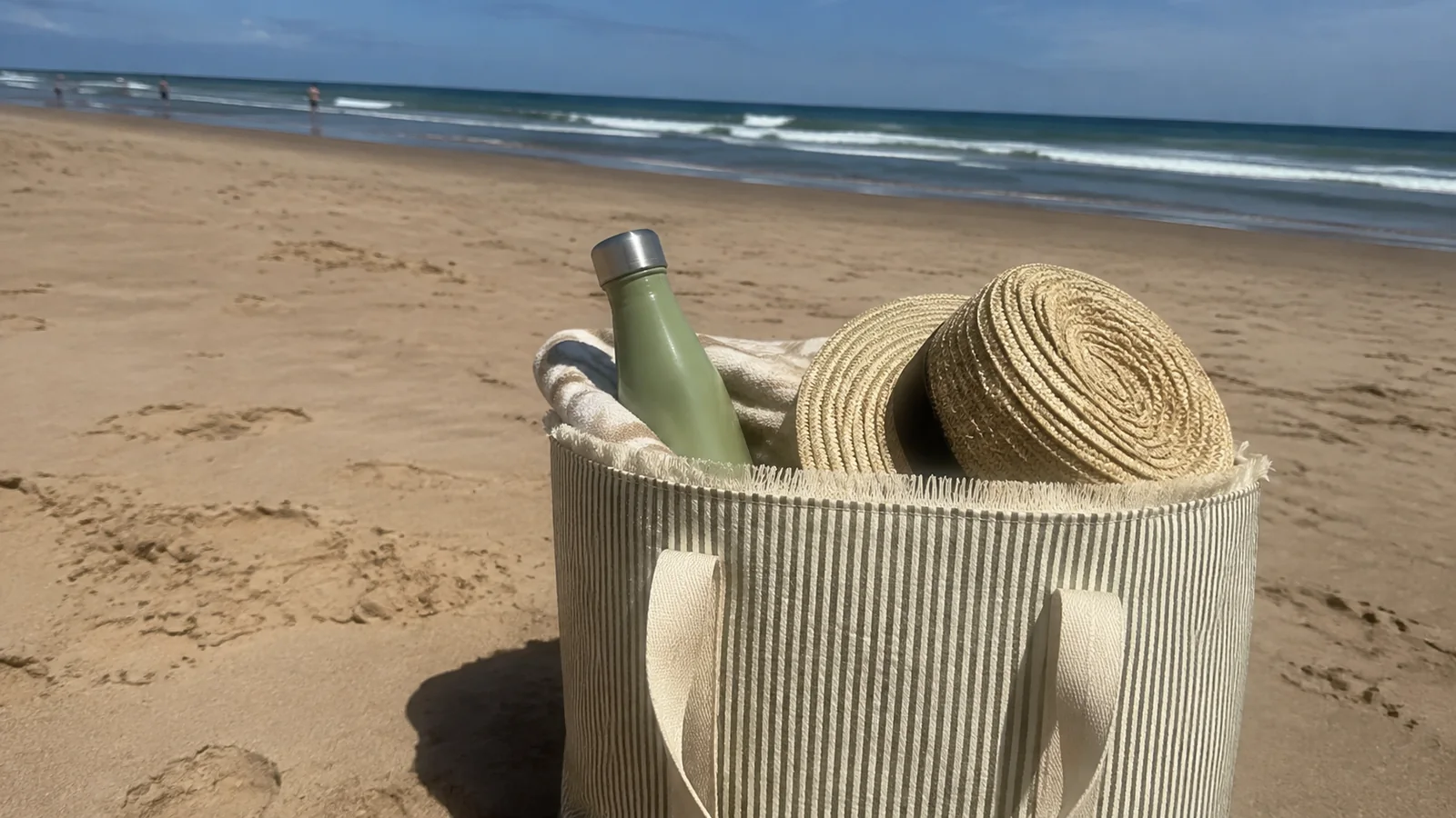 Straw hat and reusable bottle sit inside a beach tote on sand.