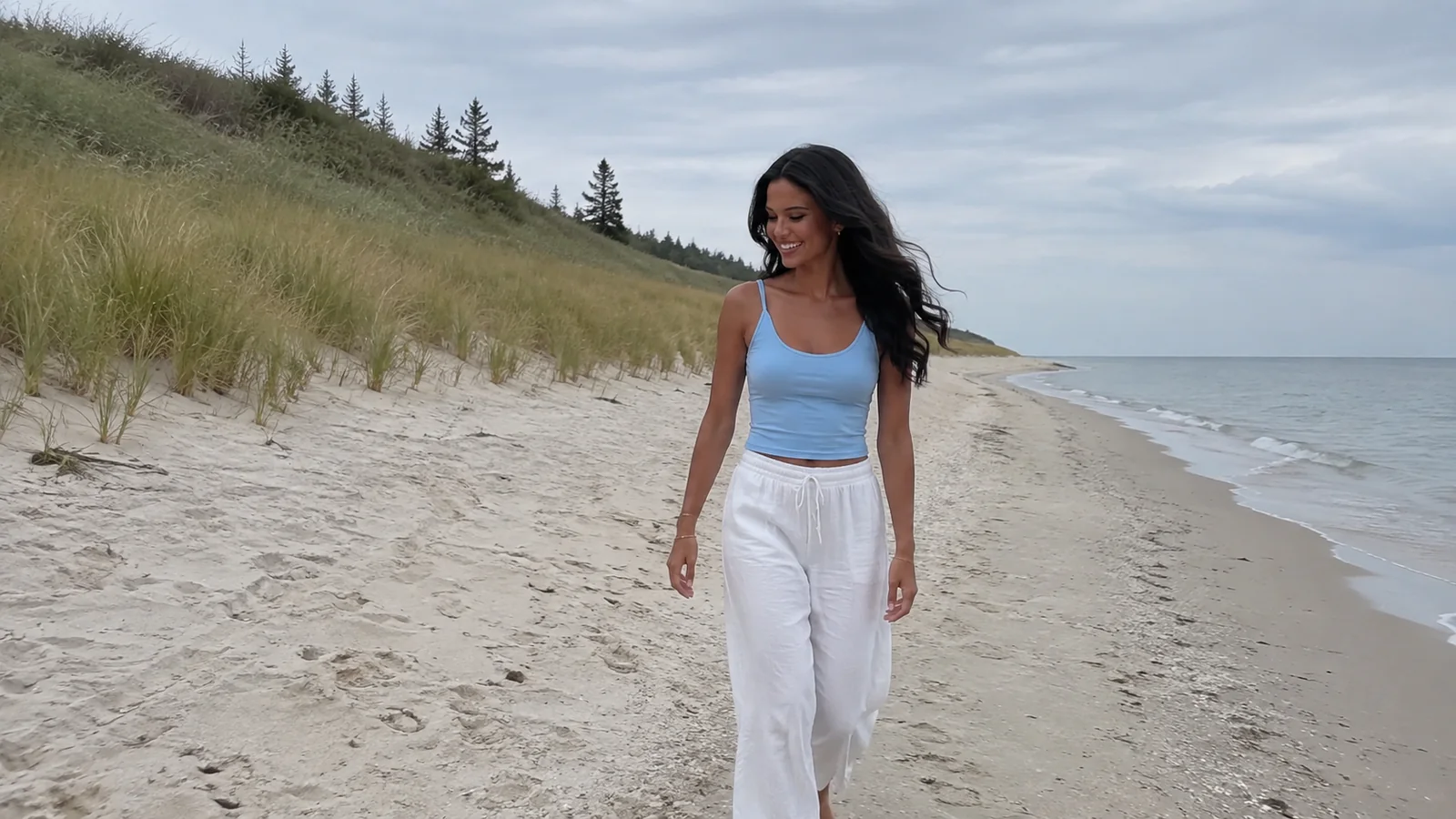 Woman walking beside pale sand dunes and gentle shore waves.