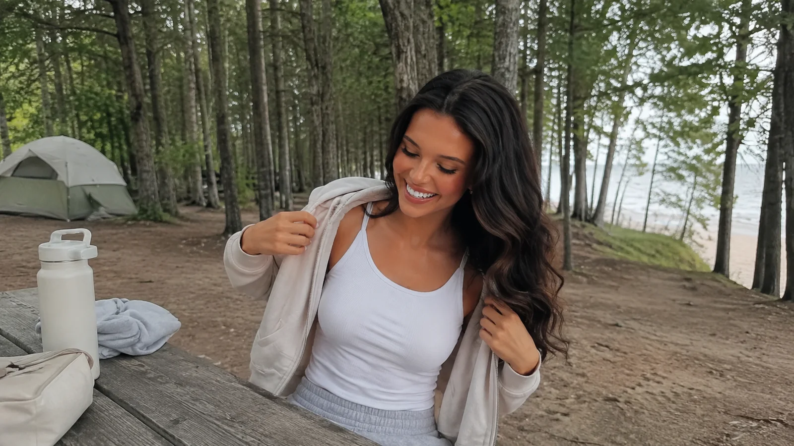 Woman adjusting her light jacket beside a wooded waterfront campsite.