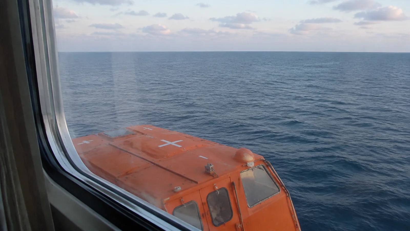 An orange lifeboat partially blocks the view through a ship window.