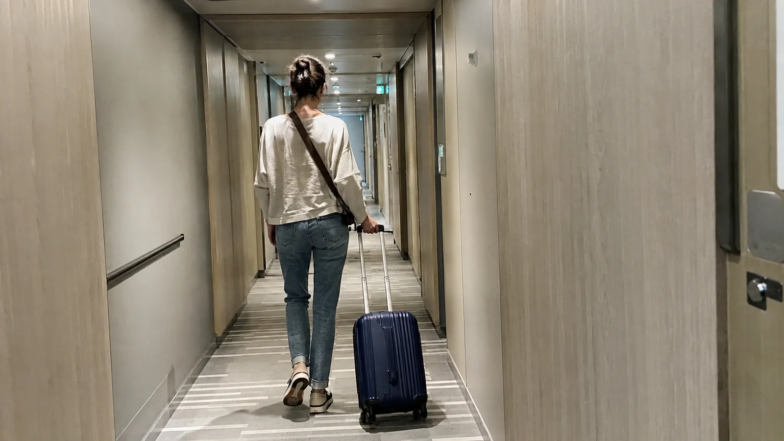Woman pulls a small suitcase along a pale ship corridor.