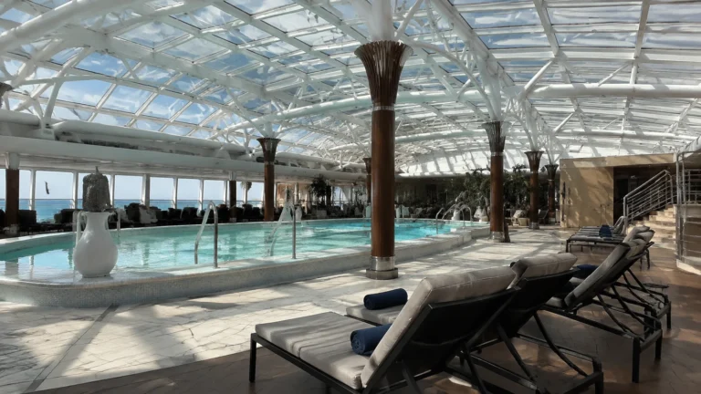 Indoor pool and loungers beneath a curved glass roof on a cruise ship.