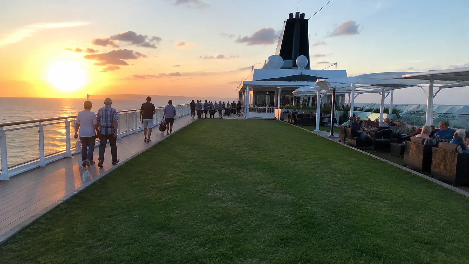 Passengers walking beside a lawn on a cruise deck at sunset.