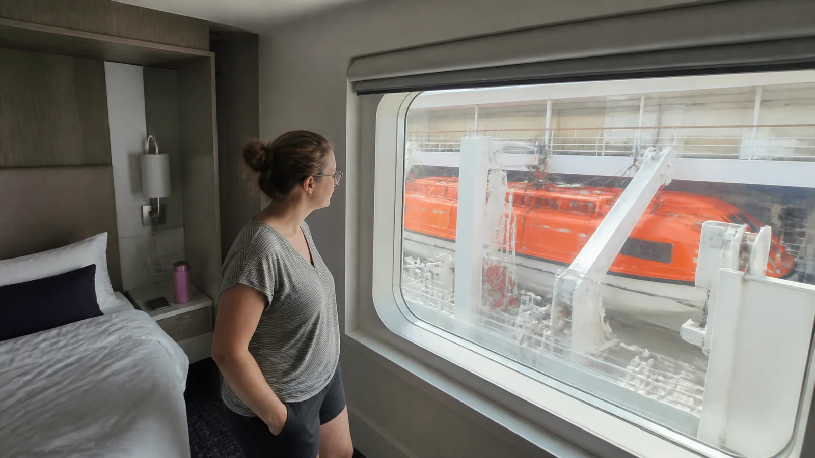 Passenger looking through a cabin window partly blocked by an orange lifeboat.