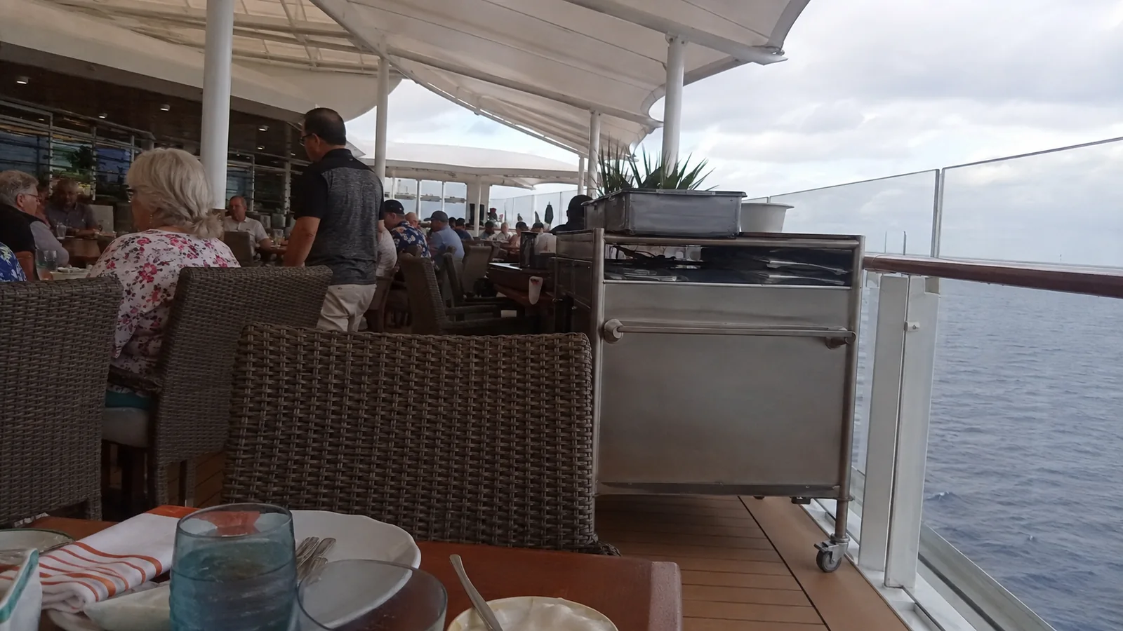 Passengers dining outdoors on a covered ship terrace.