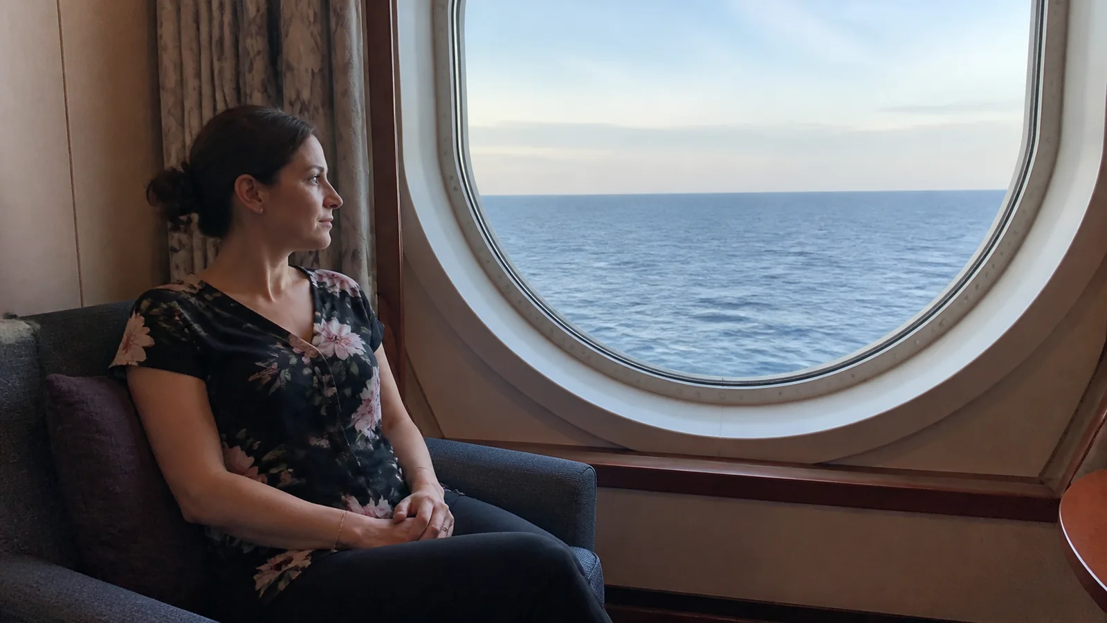 Woman sits beside a round ship window overlooking the sea.