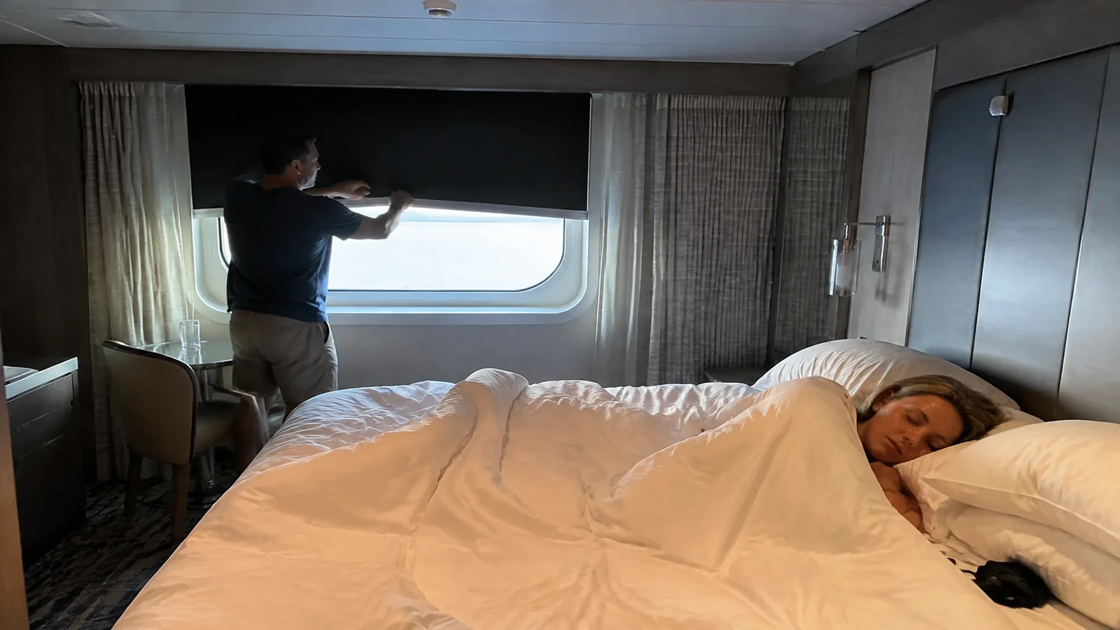 Man adjusts a window shade while a woman rests in bed.