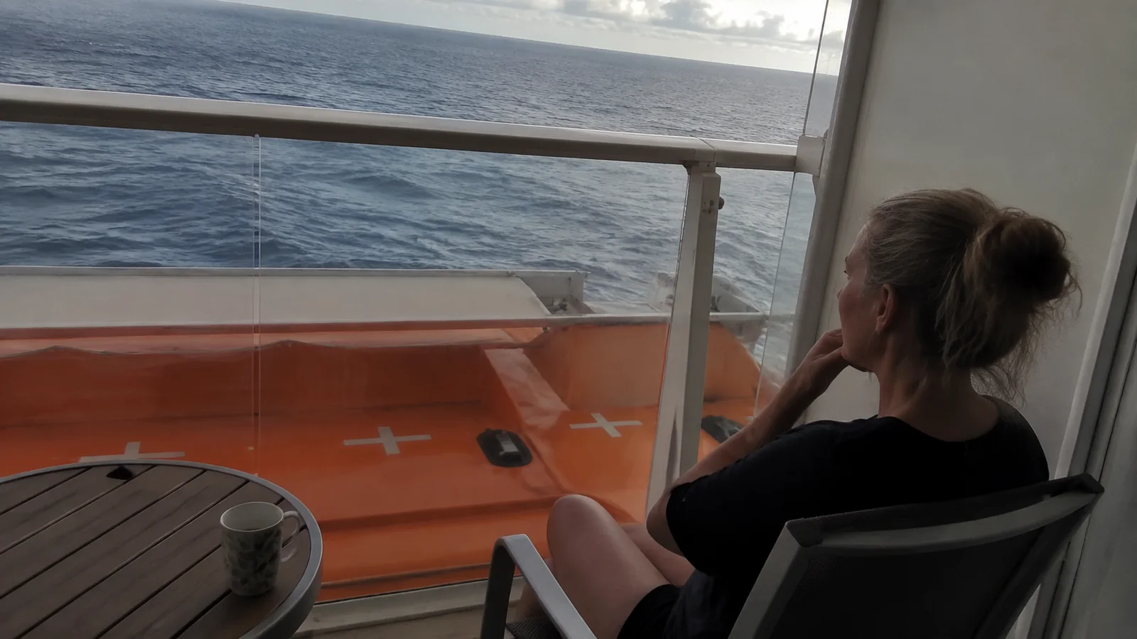 Woman sits on a ship balcony above an orange lifeboat roof.