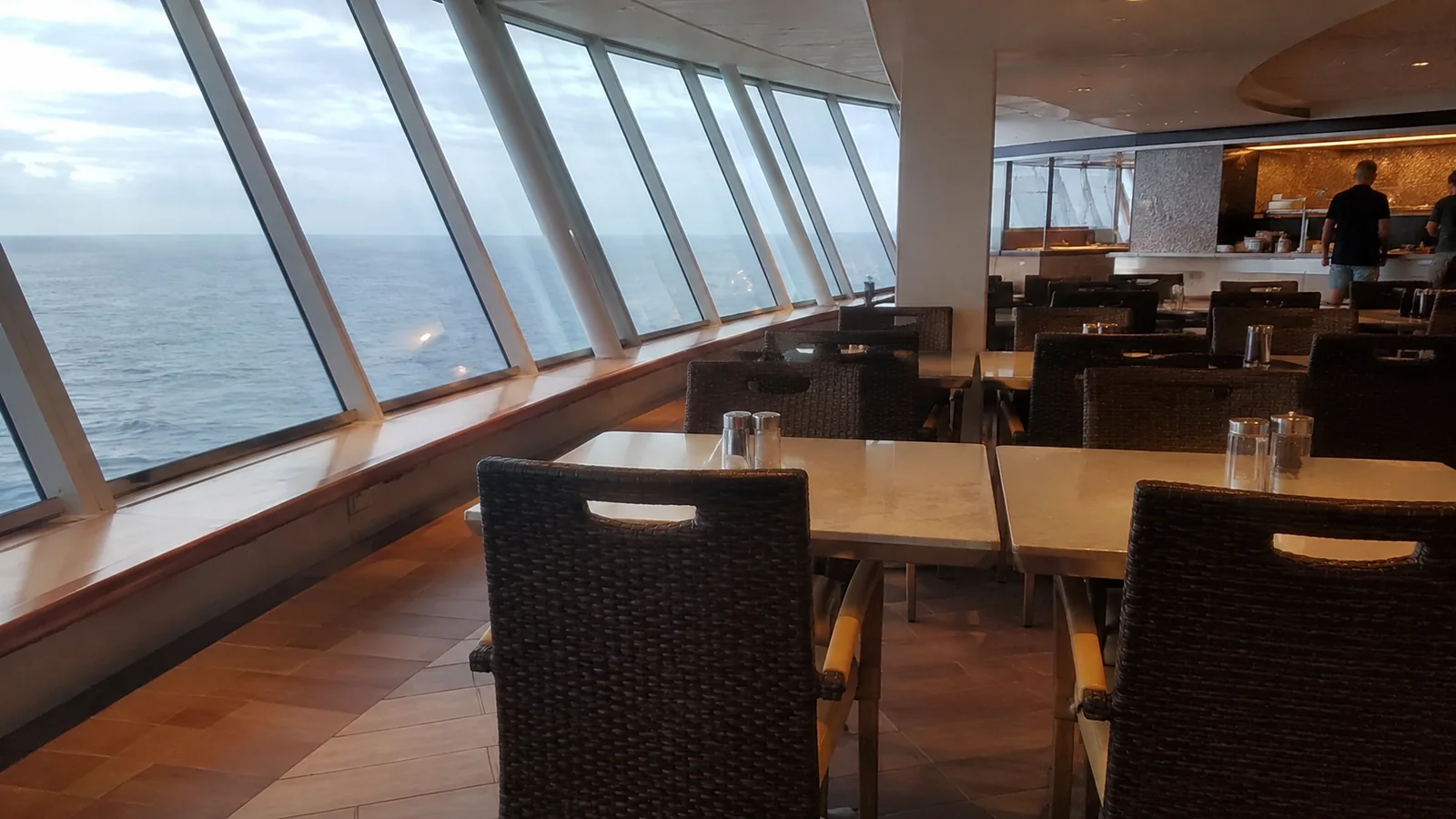 Empty ship dining tables line a row of slanted ocean windows.