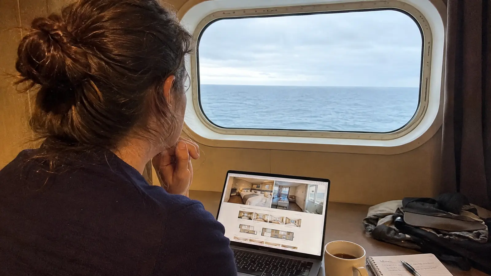 Woman using a laptop beside a large ship window overlooking the ocean